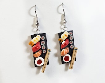 Puede incluir: Un par de pendientes colgantes con bandejas de sushi rectangulares negras. Cada bandeja muestra rollos de sushi en miniatura, piezas de nigiri, un pequeño cuenco de salsa roja y un palillo de color canela. Ganchos de metal plateado.