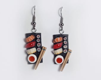 May include: A pair of dangle earrings featuring miniature sushi platters on black rectangular bases. Each earring displays salmon, tuna, and white fish nigiri, small maki rolls, a red sauce dish, and a pair of tan chopsticks with silver hooks.