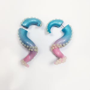 May include: A pair of unique, handmade earrings shaped like octopus tentacles. The earrings feature a gradient of colors, transitioning from blue to purple to pink. Small, white, bead-like details line the edges.