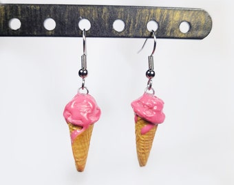 May include: A pair of dangle earrings shaped like ice cream cones. Each earring has a silver-coloured hook and a small, round bead. The ice cream cones have a waffle cone base topped with pink ice cream.