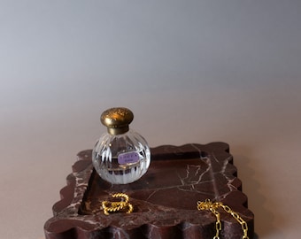 May include: A brown marble tray with a scalloped edge, featuring a clear glass bottle of perfume with a gold cap and a gold label that reads "Tocca Florence". Two gold rings and a gold chain are also on the tray.