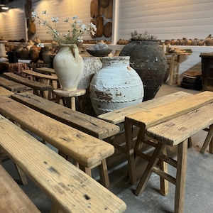 May include: A collection of rustic wooden benches with a whitewashed finish. The benches are arranged in a row, with a large white ceramic pot and a smaller white ceramic pot with flowers in the foreground. The benches are in a shop setting with a variety of other items in the background.