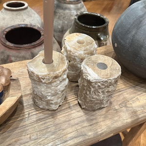 May include: Three natural stone candle holders in a rustic setting. The holders are made of a light brown stone with a rough texture. They are arranged in a row on a wooden surface.