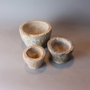 May include: Three stone bowls of varying sizes, all with a rough, textured surface. The bowls are a light beige color and are arranged in a row on a gray background.