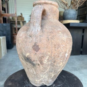 May include: A large, antique terracotta jug with a single handle. The jug has a rounded body and a wide mouth. The exterior has a weathered, textured appearance with variations in color from light brown to dark brown.