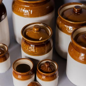May include: A collection of ceramic jars in various sizes, featuring a two-tone design with a white base and brown top. Some jars have lids, and some have text on them. The jars are arranged on a gray surface.