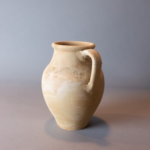 May include: A light brown clay jug with a single handle. The jug has a rounded body and a slightly flared lip.