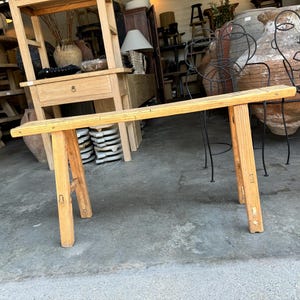 May include: A rustic wooden bench with a natural finish and a simple design. The bench has a long, rectangular top and two sturdy legs on each side. The bench is made of solid wood and has a natural, weathered look.