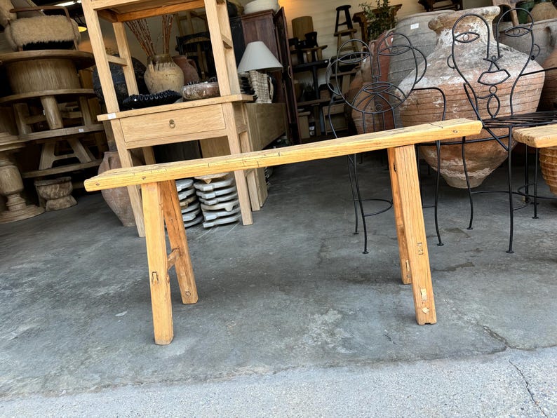 May include: A rustic wooden bench with a natural finish and a simple design. The bench has a long, rectangular top and two sturdy legs on each side. The bench is made of solid wood and has a natural, weathered look.