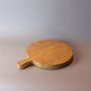 May include: A round wooden cutting board with a handle. The board is made of light brown wood and has a natural finish.