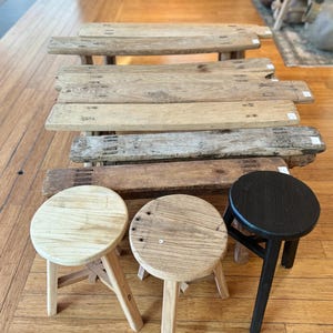 May include: Four wooden stools, two with natural wood finishes and one with a black finish. The stools have three legs and round seats. There are also three wooden benches in the background.