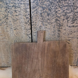 May include: A rectangular, rustic wooden cutting board with a handle. The board is a warm brown color with visible wood grain and a slightly curved edge. The background is a weathered wood panel.