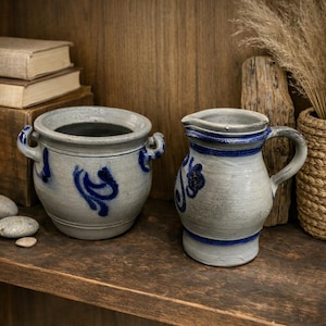 May include: Two ceramic pieces: a small, round pot with handles and a pitcher. Both are gray with blue floral designs and trim. They sit on a wooden shelf, suggesting a rustic or farmhouse aesthetic.