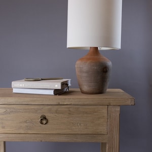 May include: A wooden side table with a single drawer and a lamp with a white shade and a brown ceramic base. A book titled "Home Body" is on top of the table.