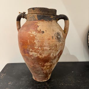 May include: A weathered, antique terracotta vase with two handles. The vase has a bulbous body and a dark rim. The exterior shows a mix of orange, brown, and beige tones, with areas of paint loss, giving it a rustic appearance.