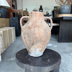 May include: A large, weathered terracotta vase with two handles. The vase has a bulbous body and a textured, aged appearance, with shades of brown and hints of blue. It sits on a dark wooden stool, showcasing its rustic charm.