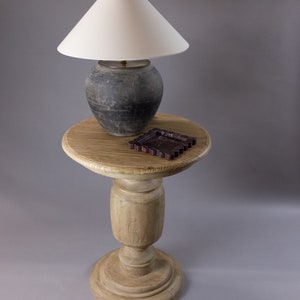 May include: A wooden pedestal table with a round top and a turned leg. A white lampshade with a dark gray ceramic base sits on the table, along with a small, dark brown decorative object.