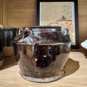 May include: A dark brown, glazed ceramic jar with two handles. The jar has a rounded body and a slightly flared base. The glaze has a glossy finish. The jar is sitting on a wooden surface.
