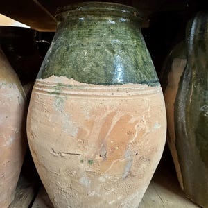 May include: A large, antique terracotta jar with a green glazed top. The lower portion of the jar is a light brown color with visible texture and imperfections. The jar has a wide body and a narrow neck, suggesting it may have been used for storage.