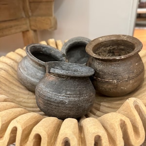 May include: Four small, dark gray clay pots with rounded bottoms and slightly flared rims sit on a light brown, carved wooden tray. The tray has a scalloped edge.