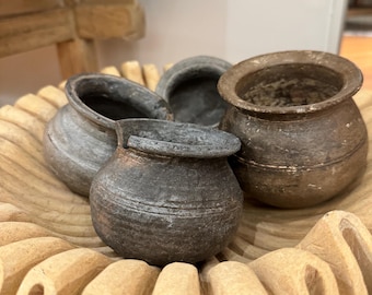 Puede incluir: Cuatro pequeñas macetas de arcilla gris oscuro con fondos redondeados y bordes ligeramente acampanados se colocan sobre una bandeja de madera tallada de color marrón claro. La bandeja tiene un borde festoneado.
