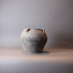 May include: A large, round, gray and white ceramic vase with two handles. The vase has a textured, painted appearance. The neutral color palette and simple design suggest a rustic or minimalist aesthetic. The vase is suitable for home decor.