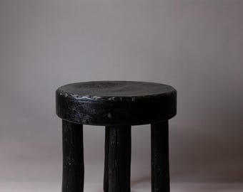 May include: A black wooden stool with a round seat and four legs. The stool is made of rough-hewn wood and has a rustic appearance.