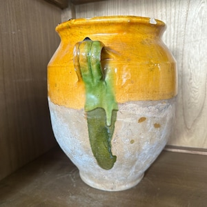 May include: A rustic, antique ceramic jar with a yellow top and a light-colored base. The jar features a green handle with a drip effect. The piece is displayed on a wooden shelf, showcasing its aged appearance and unique design.