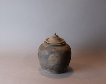 May include: A dark brown ceramic jar with a lid. The jar has a slightly rounded shape and a rough, textured surface.