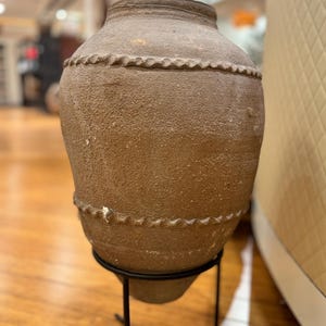 May include: A large, brown, earthenware pot with a rounded body and a narrow neck. The pot has a decorative, raised design around the middle and bottom. It is sitting on a black metal stand.