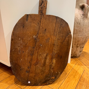 May include: A large, round, wooden bread board with a handle. The board is made of dark wood and has a rustic, antique look.
