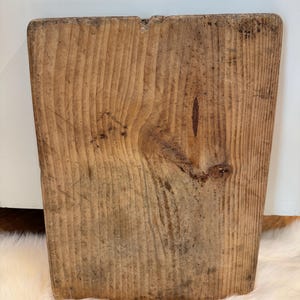 May include: A rectangular, rustic wooden cutting board with a natural wood grain pattern. The board has a slightly tapered shape and a small notch at the top. The wood has a warm, brown tone with darker knots and markings, suggesting age and use.