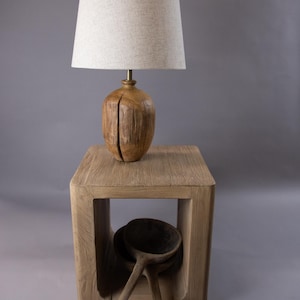 May include: A wooden lamp with a beige fabric shade sits on a wooden side table with a square open frame. Two wooden spoons are inside the frame.