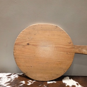 May include: A round, wooden cutting board with a handle. The board is made of light-colored wood and has a natural grain pattern. The handle is rectangular and extends from the side of the board. The cutting board is likely used for food preparation.