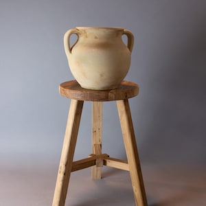 May include: A light brown clay pot with two handles sits on a wooden stool with three legs. The stool is made of natural wood with a rustic finish.