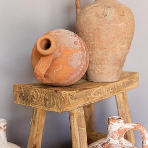 May include: Rustic terracotta jugs and a round vessel with handles sit on a weathered wooden stool. The vessels have a warm, earthy tone, with visible texture and signs of age. The stool's wood grain adds to the vintage aesthetic.