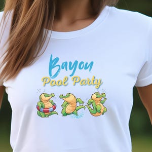 Op de afbeelding: Wit T-shirt met de tekst "Bayou Pool Party" in blauwe en gele letters. Het shirt heeft drie cartoon alligators in een zwembadfeestthema. De alligators zijn groen en geel met diverse accessoires.