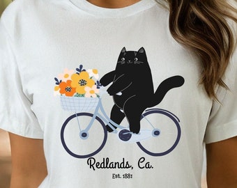 May include: White t-shirt with a black cat riding a bicycle with a basket of flowers. The text "Redlands, Ca. Est. 1881" is printed below the cat.