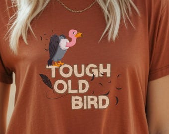 May include: A woman wearing a brown t-shirt with a pink and grey cartoon bird and the text "Tough Old Bird". She is wearing a beige hat and white jeans.