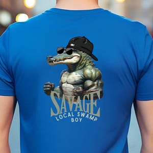 Può includere: T-shirt blu a maniche corte con una grafica di un alligatore muscoloso con occhiali da sole e cappello. Il testo "SAVAGE LOCAL SWAMP BOY" è stampato sotto l'alligatore.