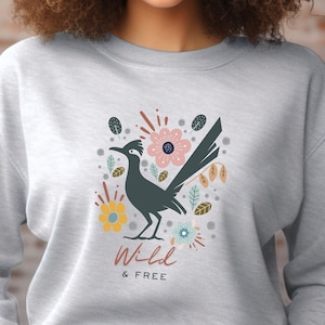 May include: A grey sweatshirt featuring a roadrunner bird graphic surrounded by flowers. The text "Wild & FREE" is printed below the bird.
