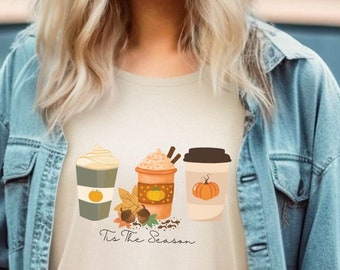 Puede incluir: Una camiseta color crema con un gráfico temático de otoño que presenta tres tazas de café con especias de calabaza y crema batida. El texto "Tis The Season" está debajo. La modelo lleva una chaqueta vaquera y vaqueros.
