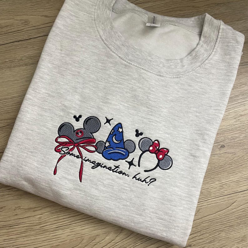 May include: Pale grey sweatshirt featuring embroidered cartoon mouse ears, a wizard's hat, and a bow. The embroidered text reads "Some imagination, huh?" below the graphics. The sweatshirt is made of a soft material.