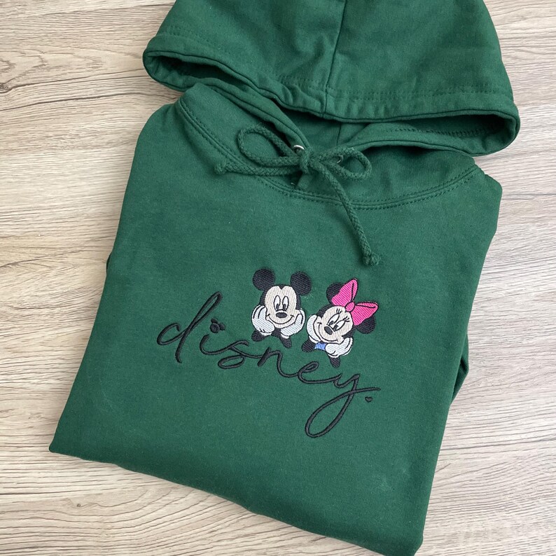 以下が含まれることがあります： 黒い刺繍でミッキーマウスとミニーマウスがデザインされた、濃い緑色のフード付きスウェットシャツ。下に「disney」の文字が刺繍されています。