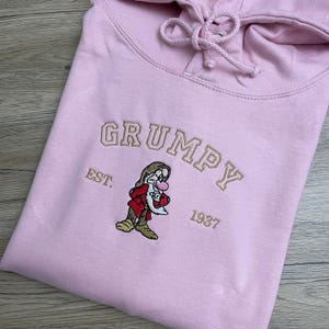 Op de afbeelding: Roze hoodie met geborduurde tekst "GRUMPY EST. 1937" en een geborduurd beeld van Grumpy uit Sneeuwwitje.