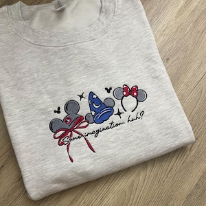 May include: A light grey sweatshirt featuring embroidered Disney designs. The design includes Mickey Mouse ears with a red bow, a blue wizard hat, and Minnie Mouse ears with a red bow. The text "Some imagination, huh?" is also embroidered.