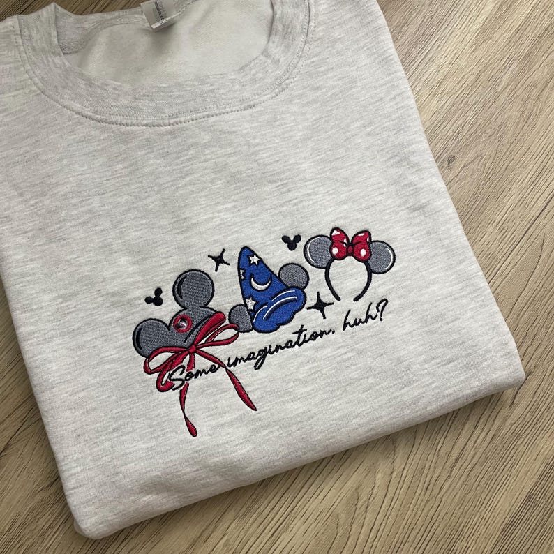 May include: A light grey sweatshirt featuring embroidered Disney designs. The design includes Mickey Mouse ears with a red bow, a blue wizard hat, and Minnie Mouse ears with a red bow. The text "Some imagination, huh?" is also embroidered.