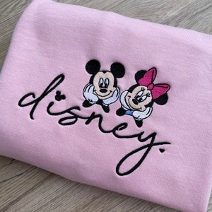 以下が含まれることがあります： ピンクのトレーナーに、黒い刺繍で「disney」という文字と、ミッキーマウスとミニーマウスのデザインが施されています。