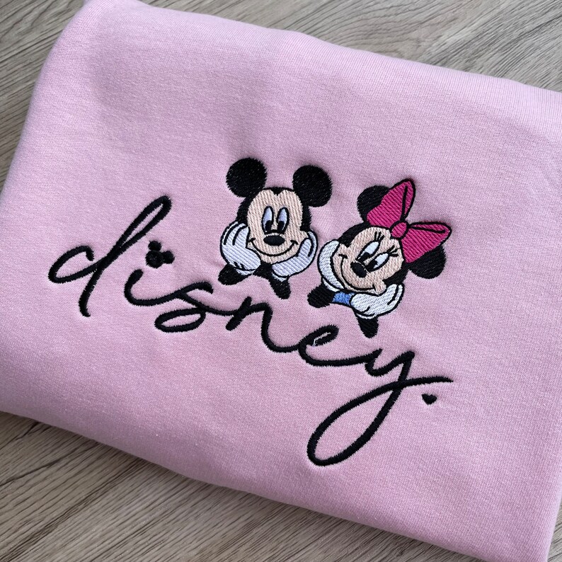 以下が含まれることがあります： ピンクのトレーナーに、黒い刺繍で「disney」という文字と、ミッキーマウスとミニーマウスのデザインが施されています。
