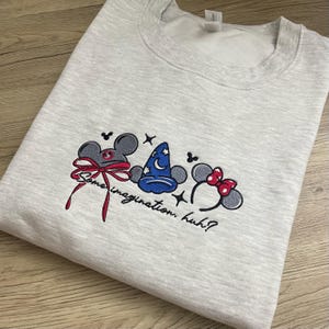 May include: Pale grey sweatshirt featuring embroidered Mickey Mouse ears, a wizard's hat, and Minnie Mouse ears. The design includes the text "Some imagination, huh?" in a script font. The sweatshirt is made of a soft material.